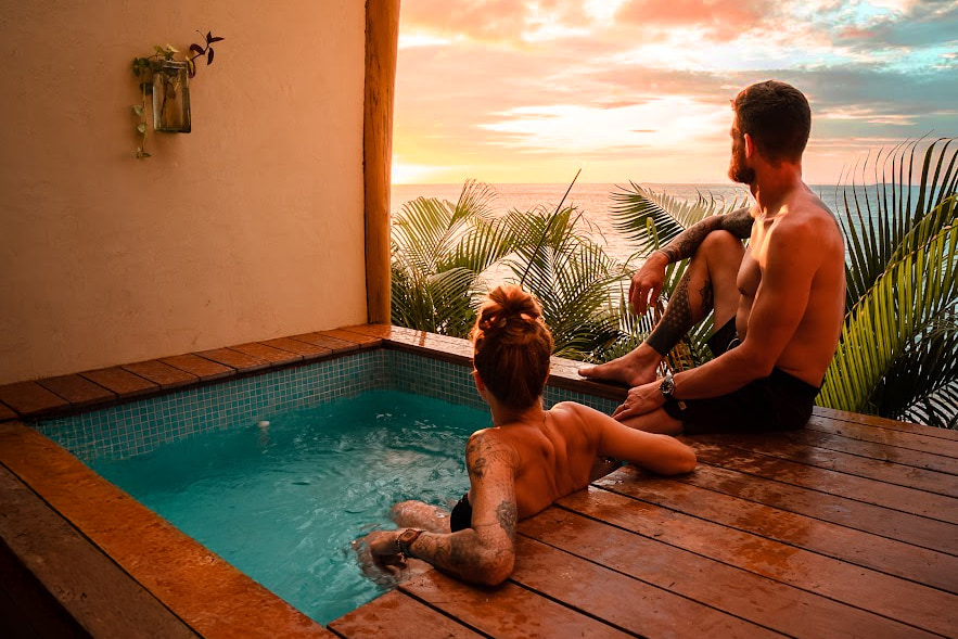 Una pareja joven y atlética está sentada en el jacuzzi de nuestra Beach Casita. Miran el atardecer y el mar a través de las hojas de las palmeras que están un nivel abajo, en la playa.