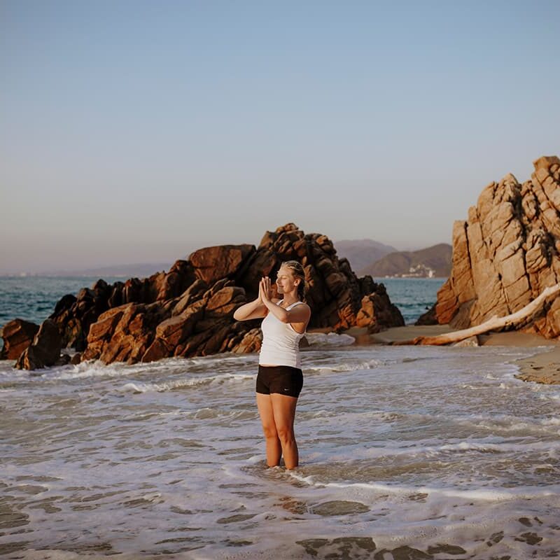 Las olas cubren los pies de una mujer en la playa de Xinalani. Ella mira en dirección al sol, con las manos en agradecimiento